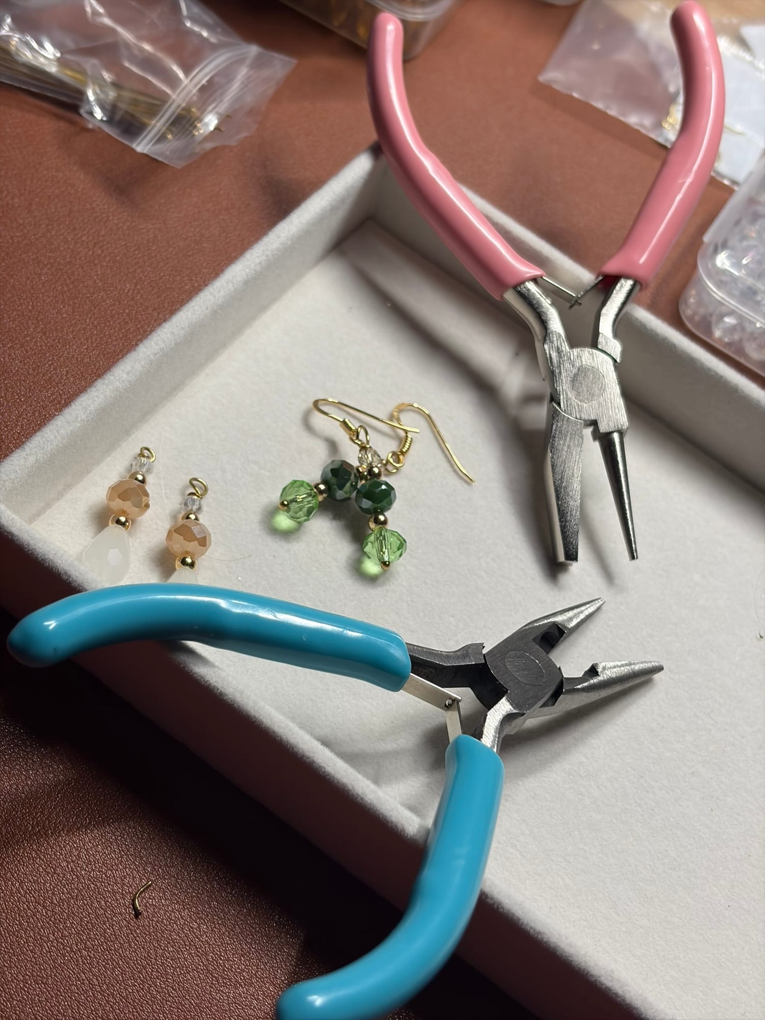 Deux pinces de bijoutier — une à poignée rose, une à poignée bleue — encadrant une boucle verte et dorée en cours de montage, avec son pendant