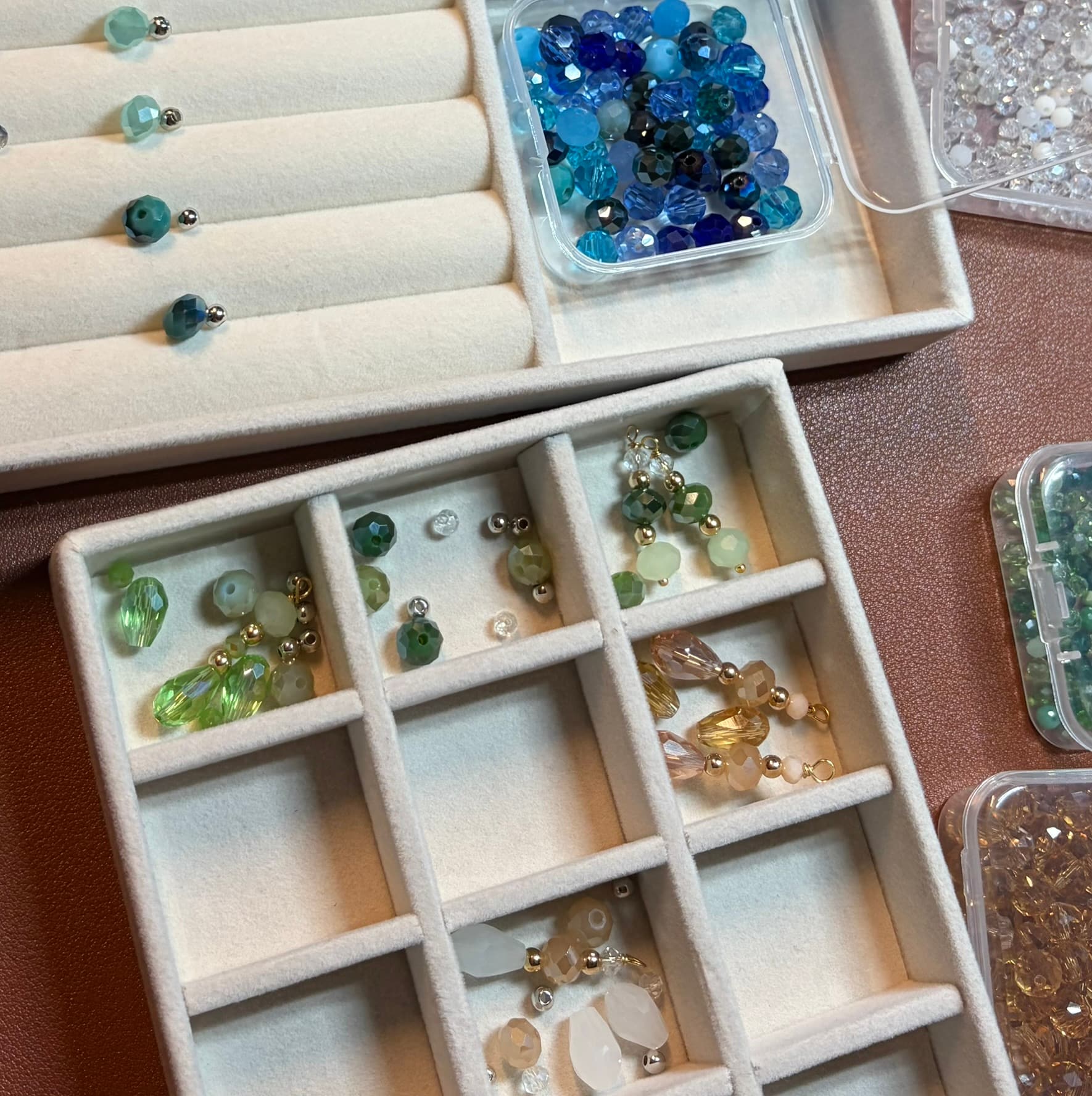 Vue en plongée du poste de travail : un plateau compartimenté de velours beige avec les perles appariées, entouré des boîtes de perles bleues, dorées et blanches